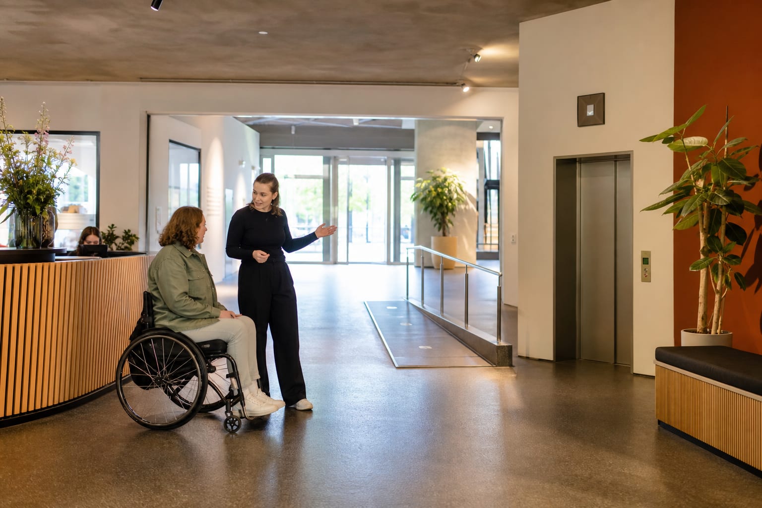 Een bezoeker in een rolstoel bespreekt met een medewerker de route langs een hellingbaan, lift en zitbank in een ruime entreehal
